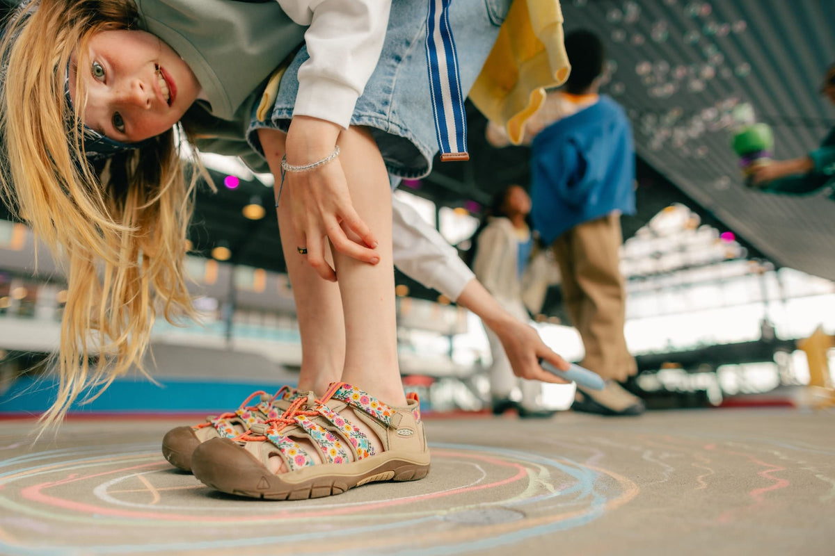 A child wearing colorful patterned slide sandals and denim shorts in a skatepark, looking down at the camera with chalk drawings on the ground.