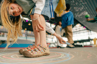 A child wearing colorful patterned slide sandals and denim shorts in a skatepark, looking down at the camera with chalk drawings on the ground.