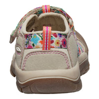 A children's casual shoe shown from the side profile on a white background. The upper is primarily beige suede with a colourful floral patterned strap and pull tab, pink stitching, and a brown rubber sole.