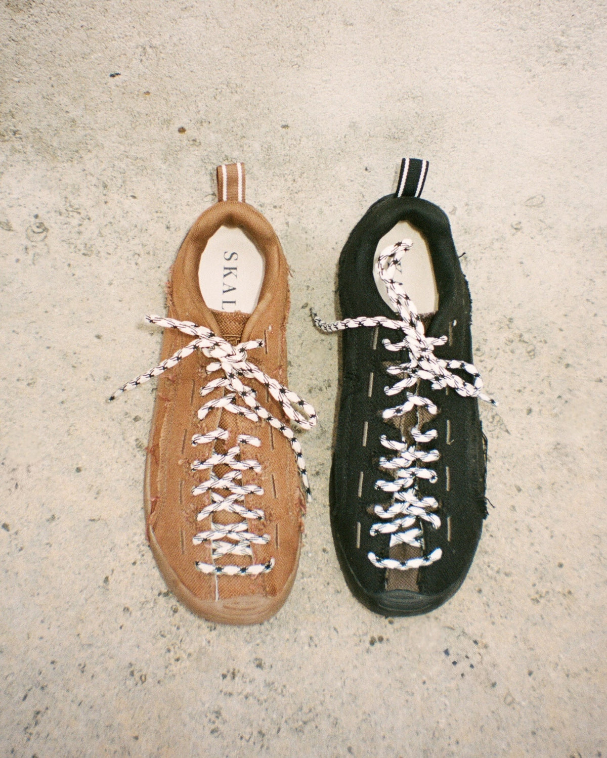 Two low-top lace-up shoes laid flat on a textured concrete surface: one tan/camel and one black, both with white-and-black braided laces. The tan shoe shows the brand name SKAI on the insole.