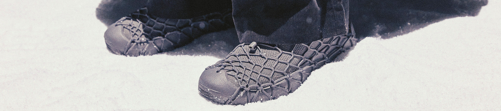 A close-up of a person's feet wearing dark gray quilted snowboard boots with integrated crampons, standing in deep snow.