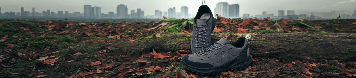 A pair of dark gray lace-up KEEN shoes placed on a grassy field scattered with fallen autumn leaves, with a blurred city skyline in the background.