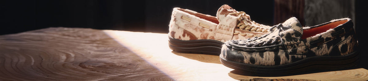 A pair of slip-on loafers with a textured woven pattern, one in beige and white and the other in black and white, displayed on a wooden surface with dramatic side lighting.