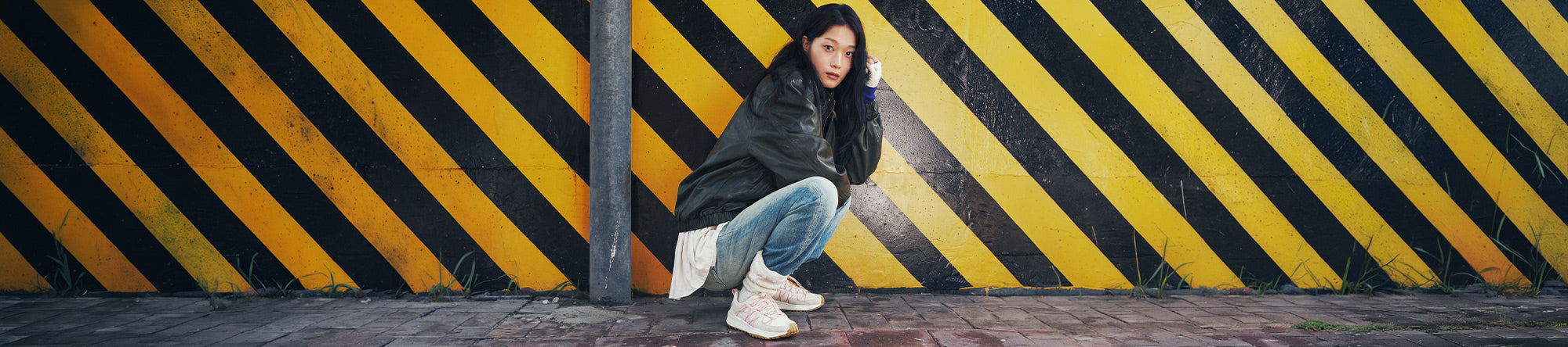 A woman crouches against a wall with bold yellow and black diagonal hazard stripes. She is wearing a dark oversized jacket, light blue jeans, and white sneakers.