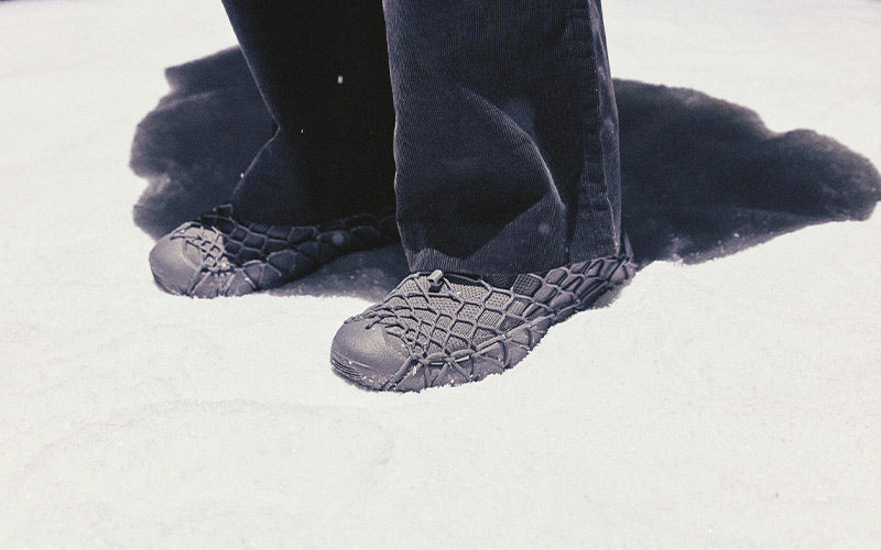 A close-up of a person's lower legs and feet wearing dark gray textured snow pants and matching snow boots standing in white snow.