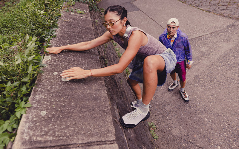 Two people on a city street, one climbing a wall and the other walking.