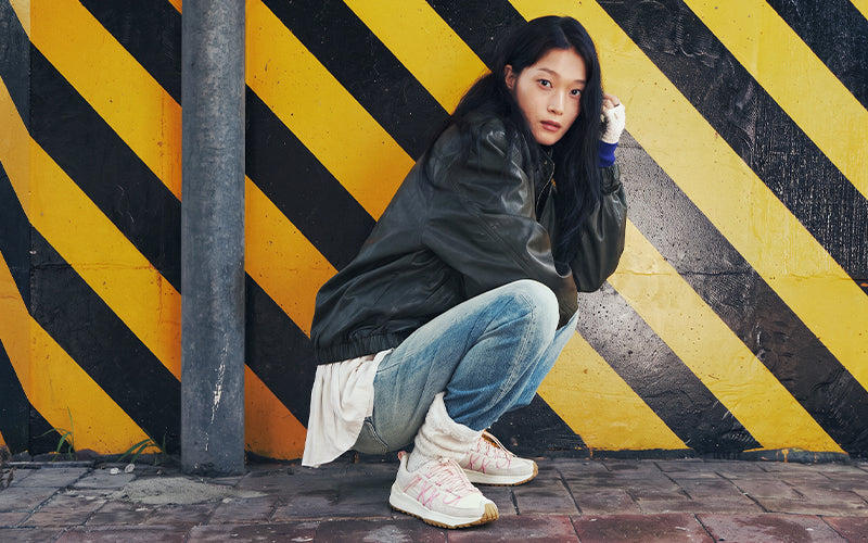 A woman crouches against a yellow and black striped wall, wearing a dark leather jacket, light blue jeans, a white shirt, and white sneakers.