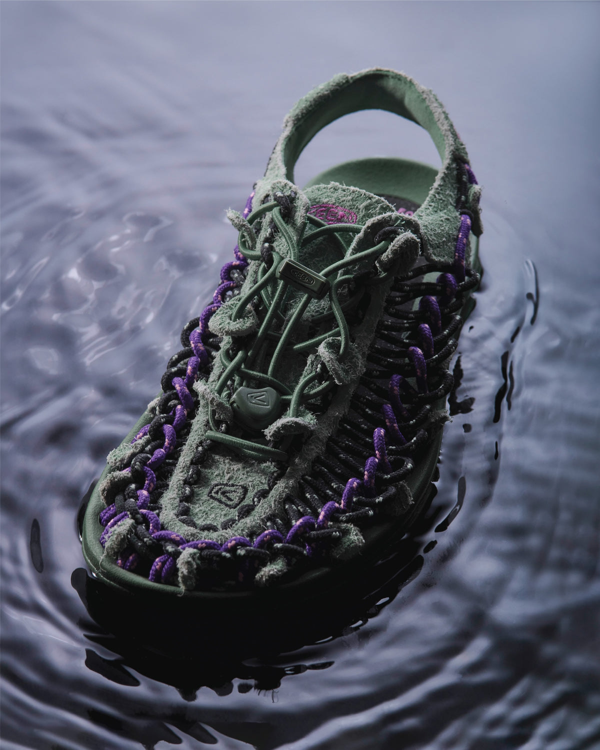 A green and purple woven outdoor sandal floating on dark water, with ripples around it.