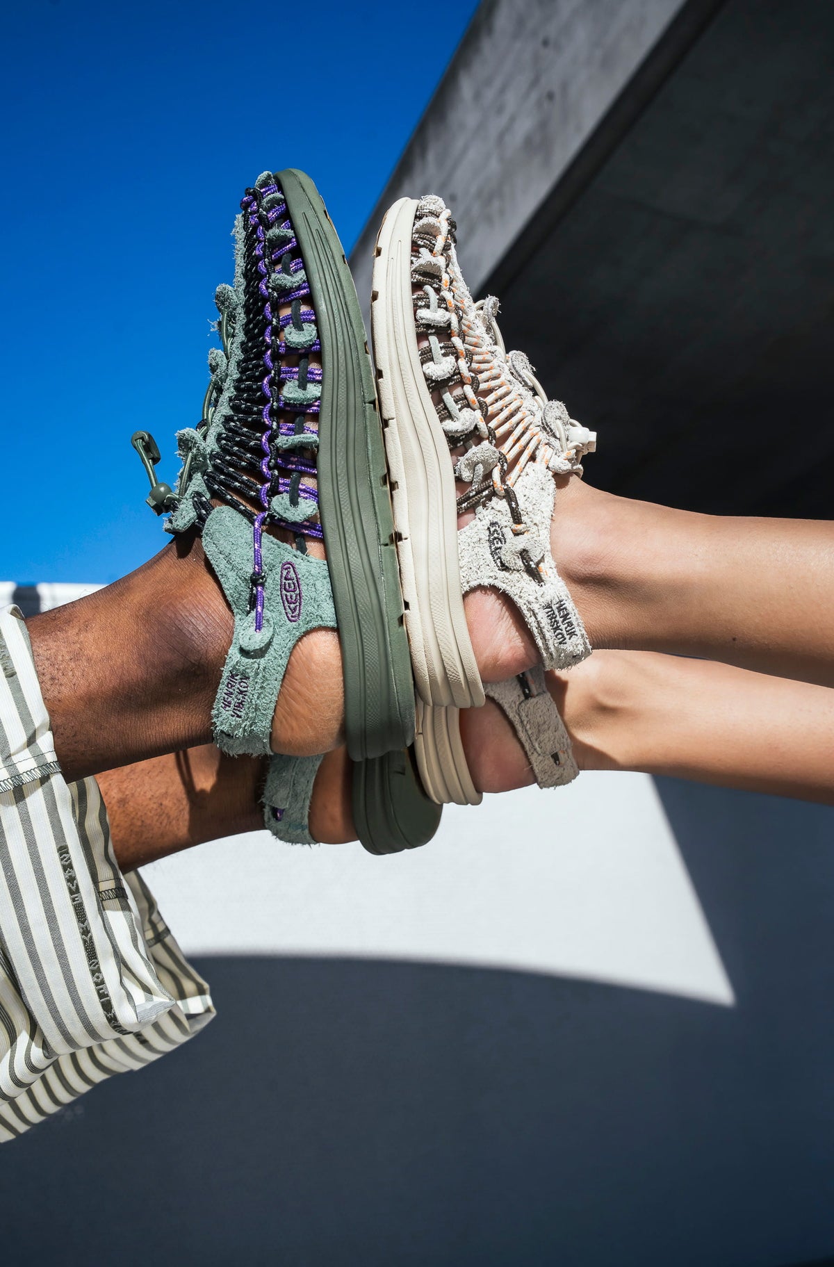 Two pairs of shoes held up against a blue sky and concrete wall.