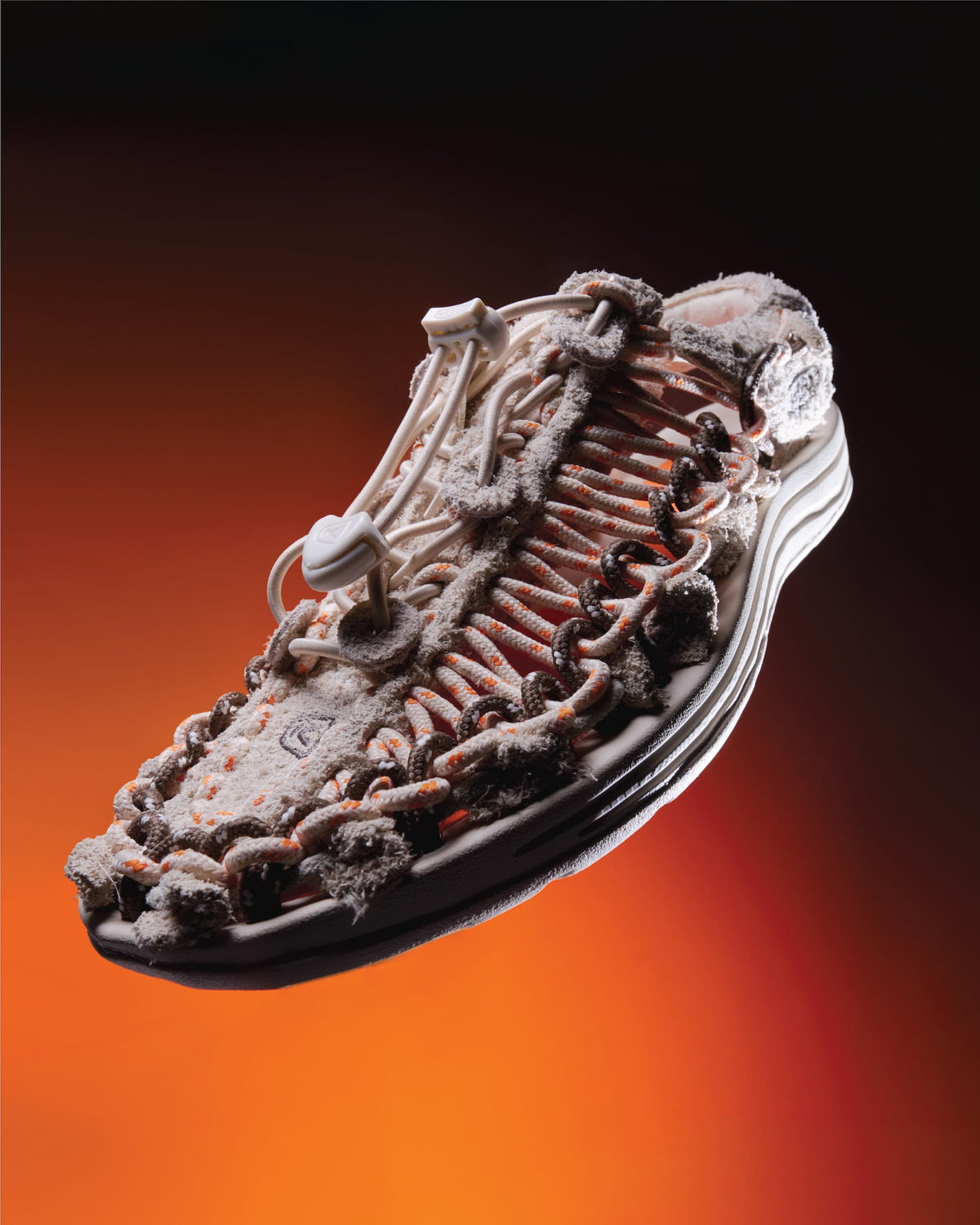 A highly detailed, textured shoe or boot shown floating against a dramatic orange-to-black gradient background. The upper is covered in white powder or dust, with dark brown and black ribbed detailing along the sole and sides.