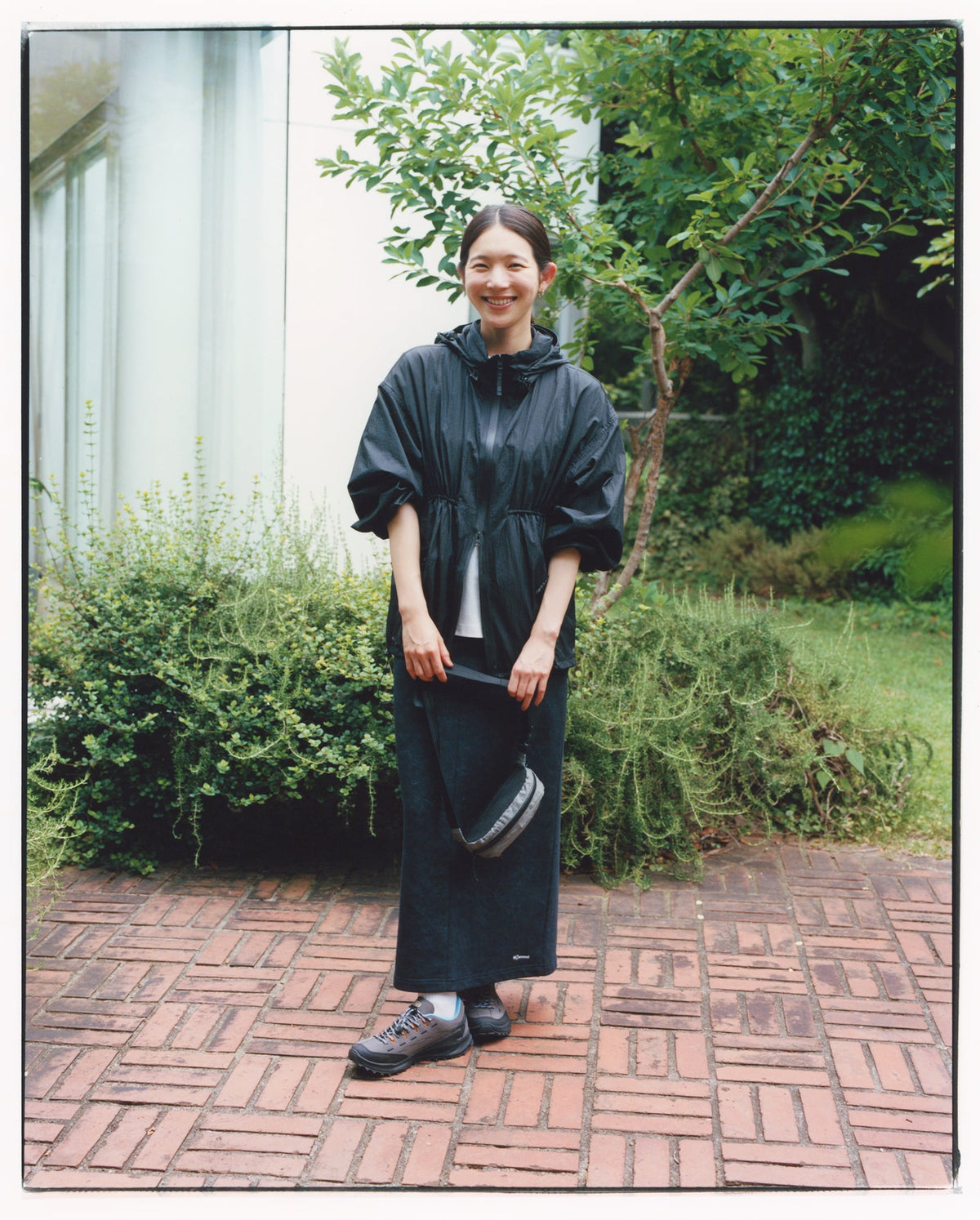 Person wearing a dark poncho standing on a brick path with greenery in the background