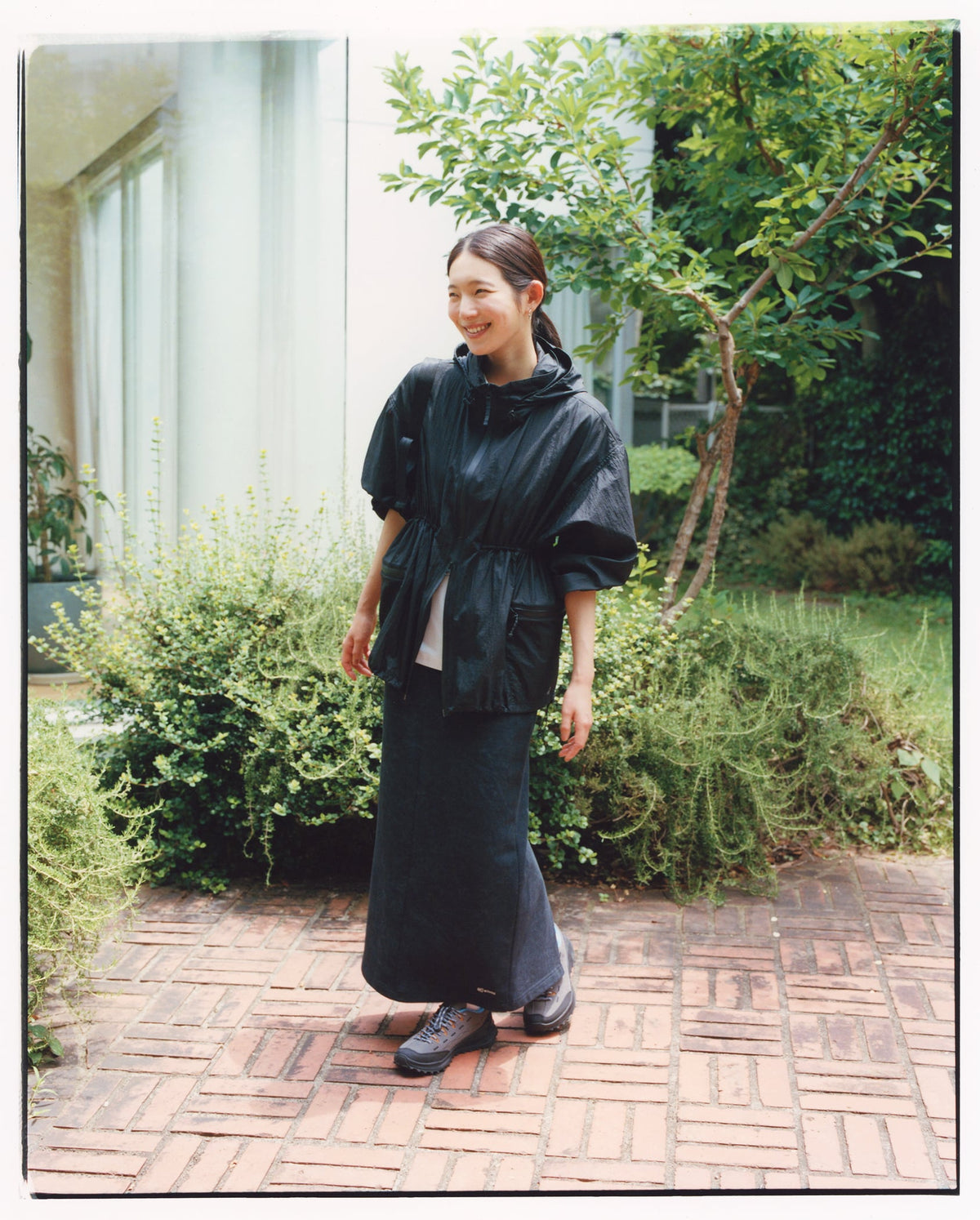 Woman in a black dress standing outdoors on a brick path with greenery around.
