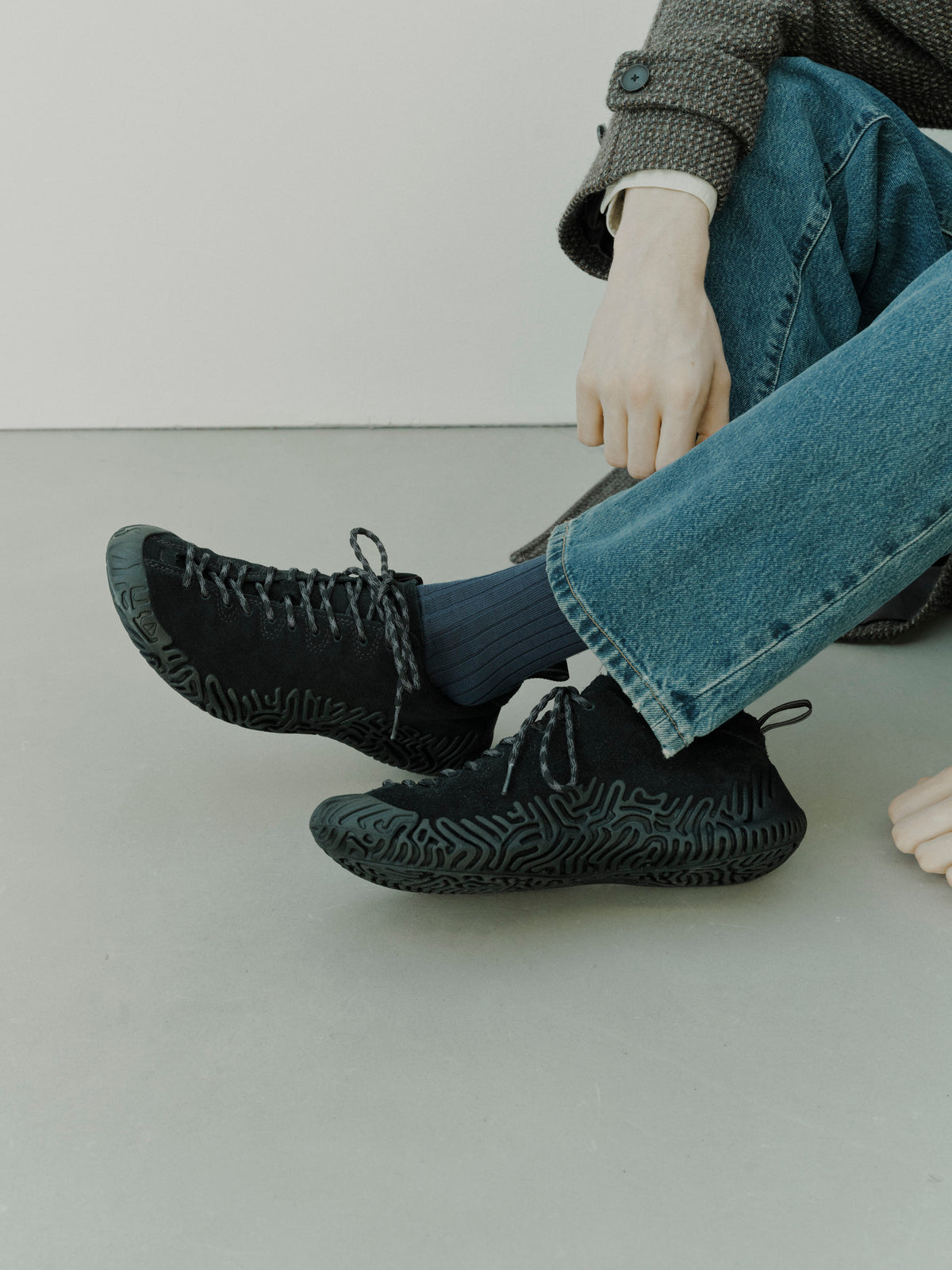 A person sitting on a light gray floor wearing black textured sneakers, blue jeans, and dark blue socks.