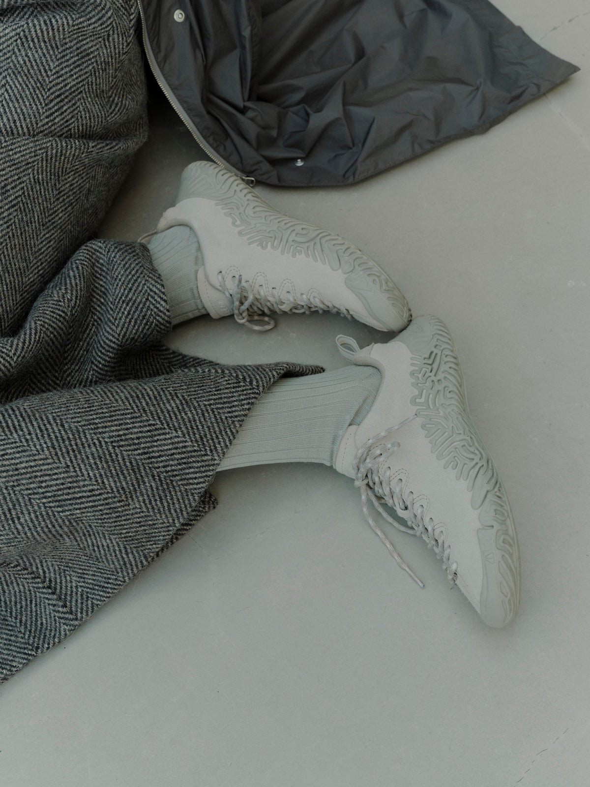 A pair of light gray and white textured sneakers with matching gray socks, styled with a gray herringbone coat and a dark gray puffer jacket on a light gray floor.