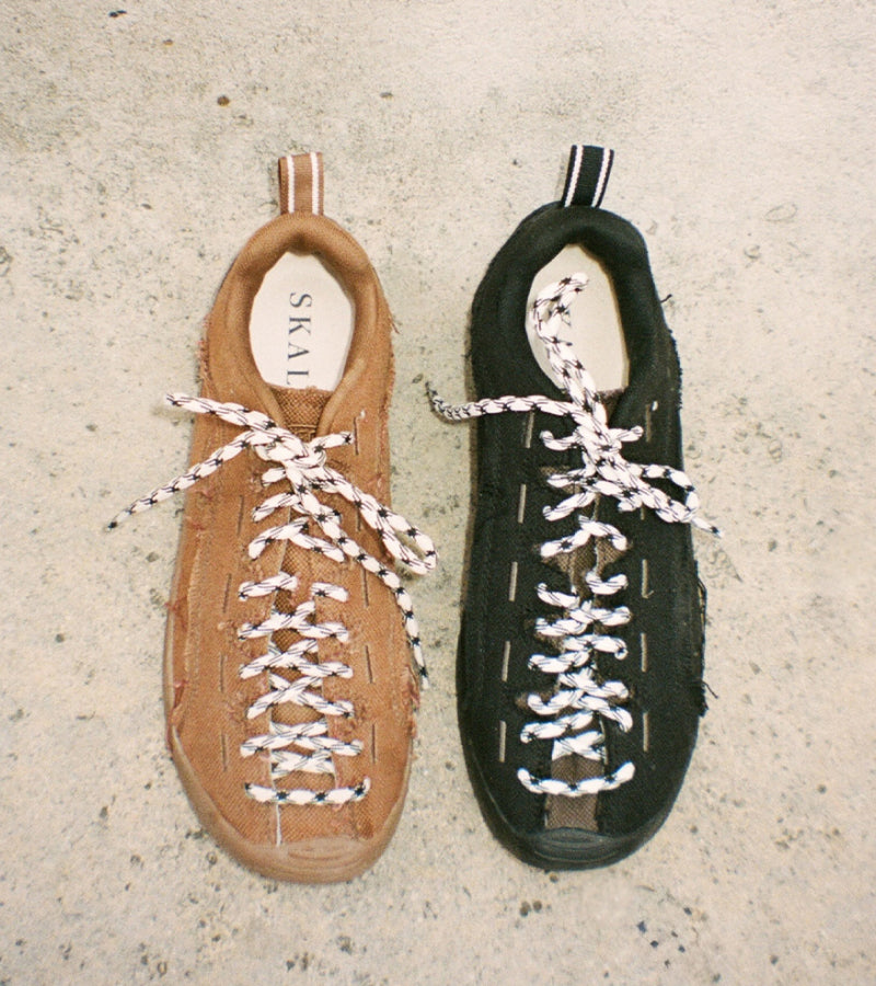 Two low-top lace-up shoes one in tan suede and one in black suede, both with white-and-black patterned laces, displayed side-by-side on a textured concrete surface.