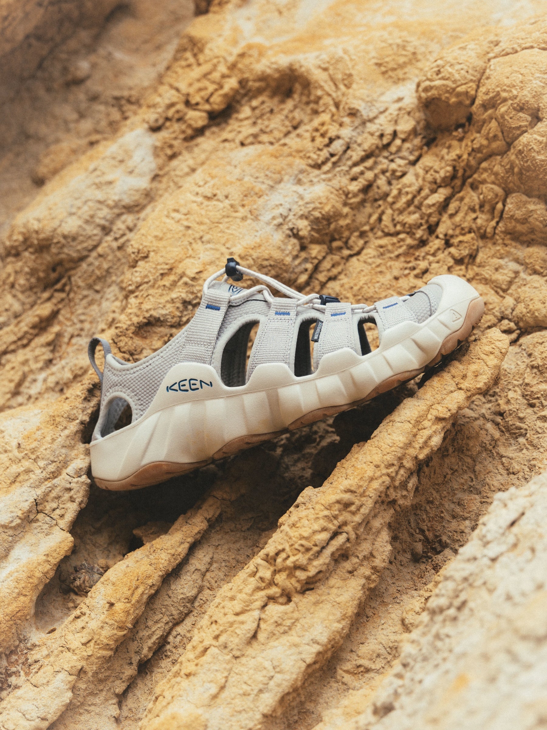 A Keen sandal in light gray and off-white, photographed resting on a textured sandy rock surface.