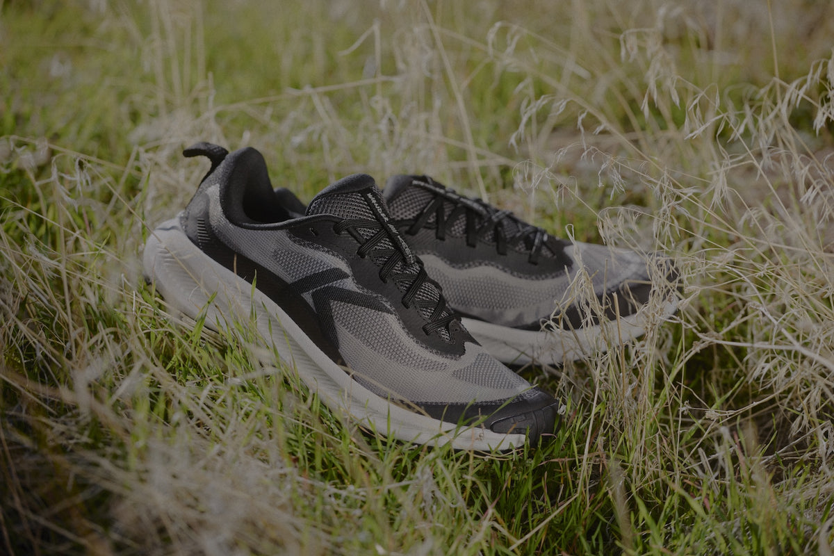 A pair of gray and black athletic trail running shoes resting on a bed of green and dry grass outdoors.