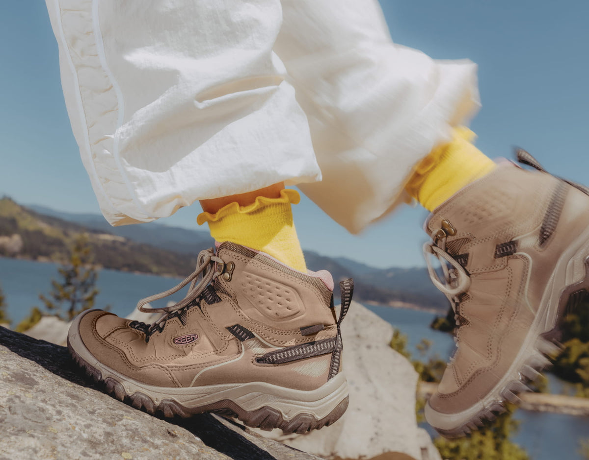A close-up of a person wearing beige hiking boots with white pants and bright yellow socks, standing on a rock with a scenic lake and mountains in the background.