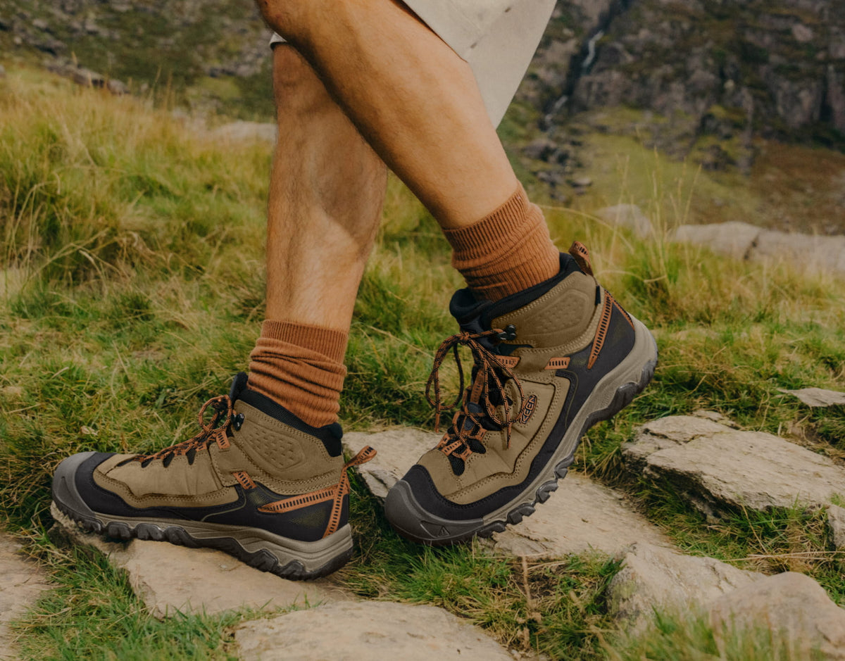A close-up of a person's legs wearing brown and black hiking boots with brown socks, standing on a grassy outdoor path with rocks and hills in the background.