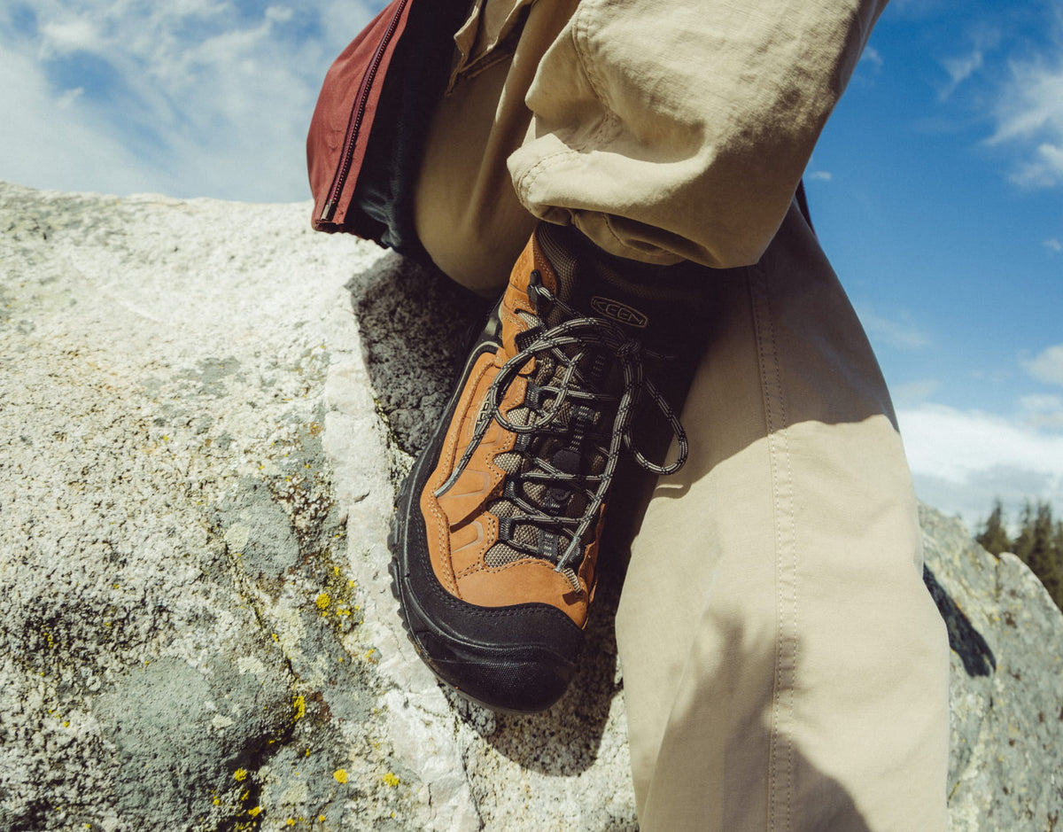 A close-up shot of a person's foot wearing a brown and black Hike-On hiking boot, paired with beige pants, sitting on a large gray rock under a blue sky.