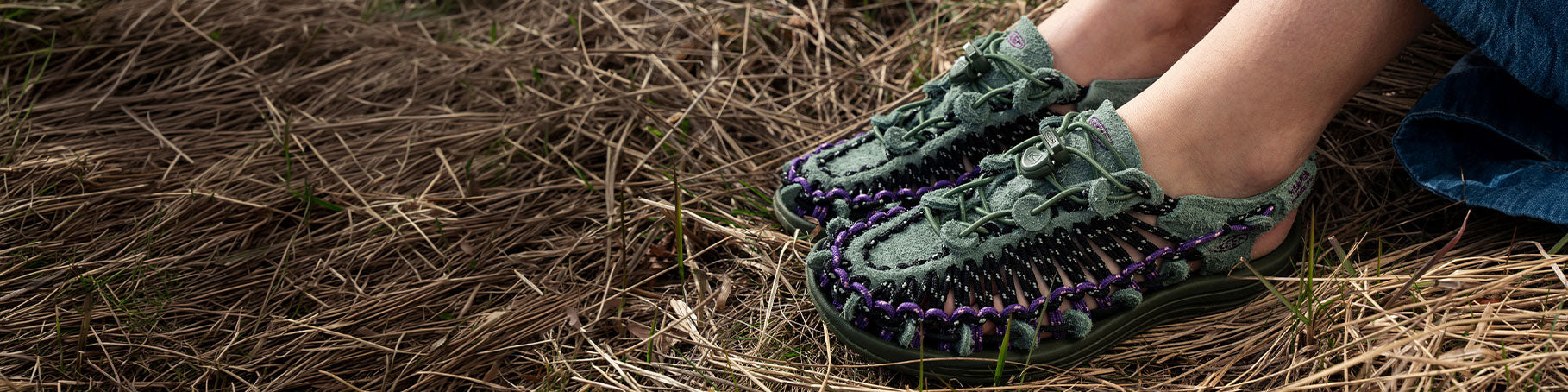 A close-up of a person's feet wearing handmade woven moccasin-style shoes outdoors on dry grass and pine needles. The shoes are olive green with purple and black braided accents around the sole.