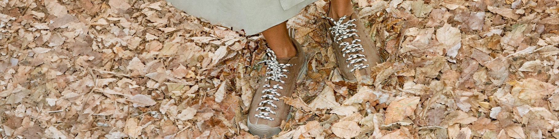 A person wearing brown suede lace-up sneakers with white laces, standing on a ground covered in dry beige and brown leaves.