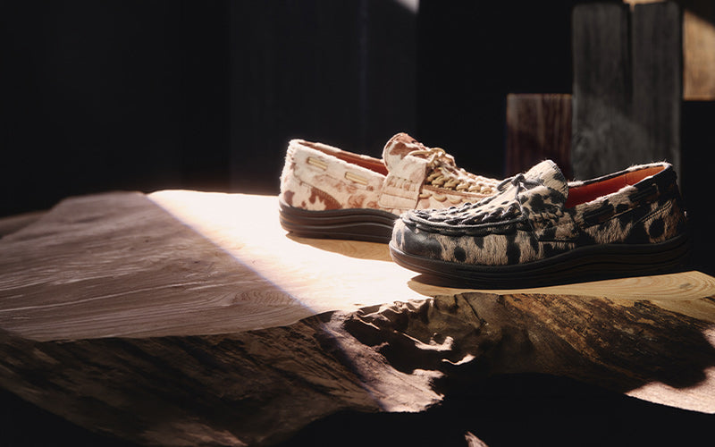 Two slip-on boat-style shoes with textured animal-print uppers, one in beige and brown and the other in black and beige, displayed on a rustic wooden slab with dramatic side lighting.