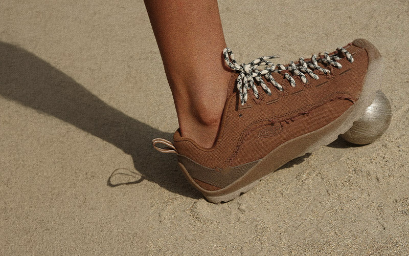 A close-up of a person's foot wearing a brown suede shoe with black and white patterned laces, standing on sand.