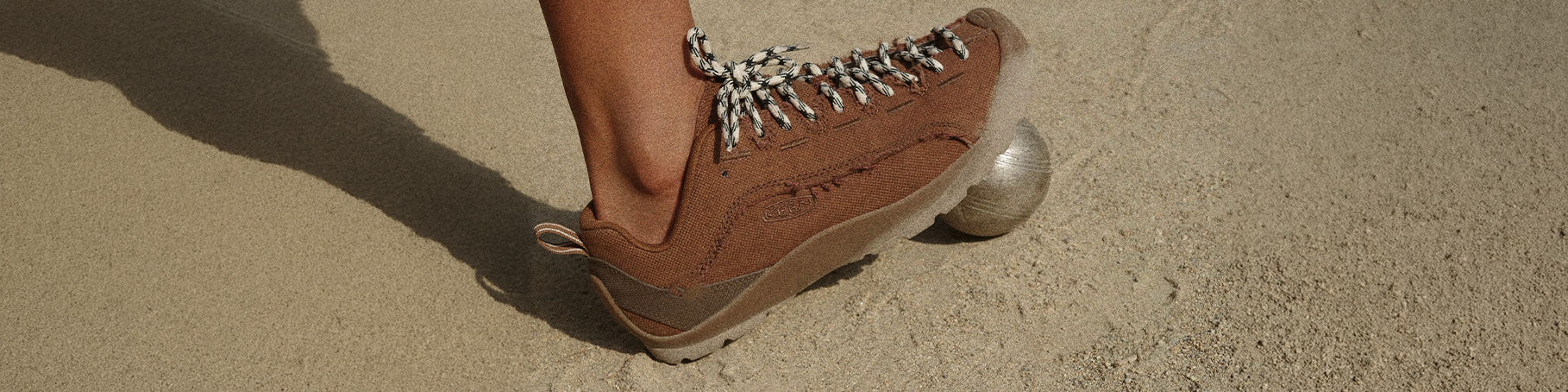 A close-up of a person's foot wearing a brown suede sneaker with silver chain laces and a metallic sole, standing on a sandy surface.