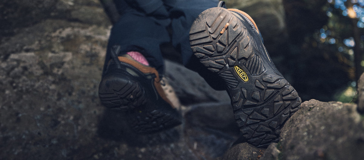 A close-up of a person's feet wearing black Keen hiking boots with brown laces, climbing over dark rocks outdoors. The sole of the raised boot is clearly visible, showing the Keen logo.
