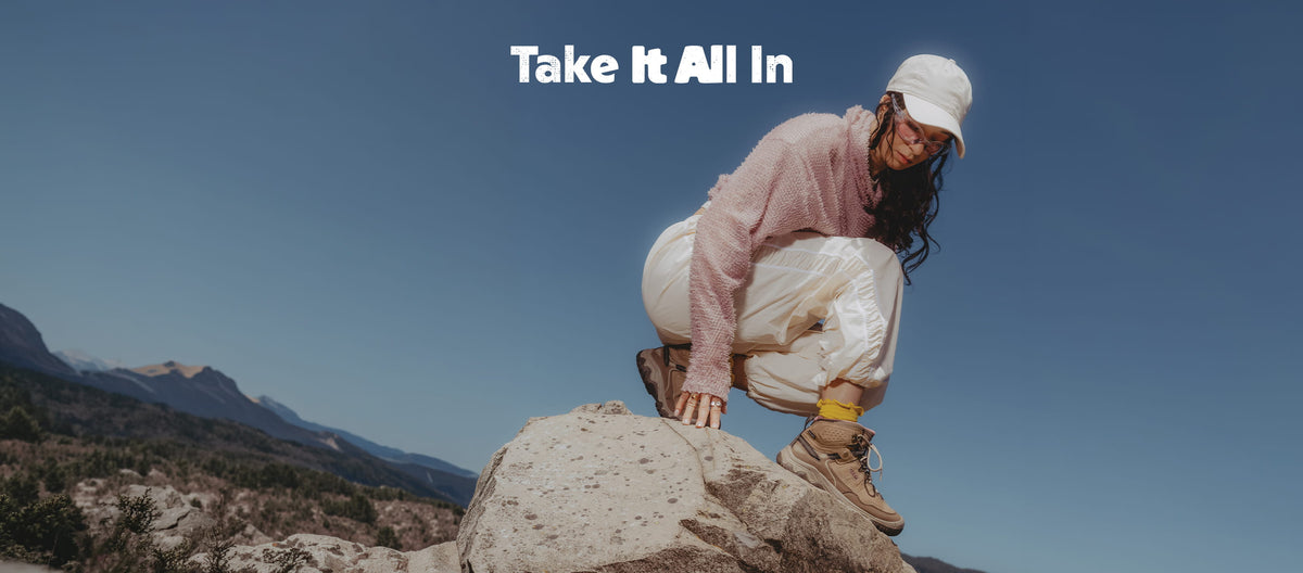 A woman crouches on a large rock in an outdoor mountain setting under a clear blue sky, wearing a light pink sweatshirt, white cap, white pants, and hiking boots. The text 'Take It All In' appears at the top.