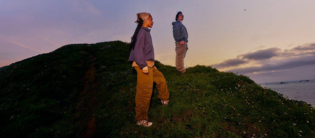 Two people standing on a grassy hillside at sunset, wearing loose outdoor hoodies and pants, with water and sky in the background.