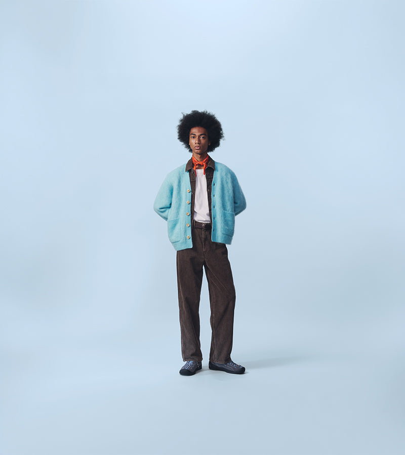 A person with an afro hairstyle stands against a light blue studio background wearing a light blue fuzzy cardigan over a white shirt with an orange neck scarf, dark brown trousers, and dark shoes.