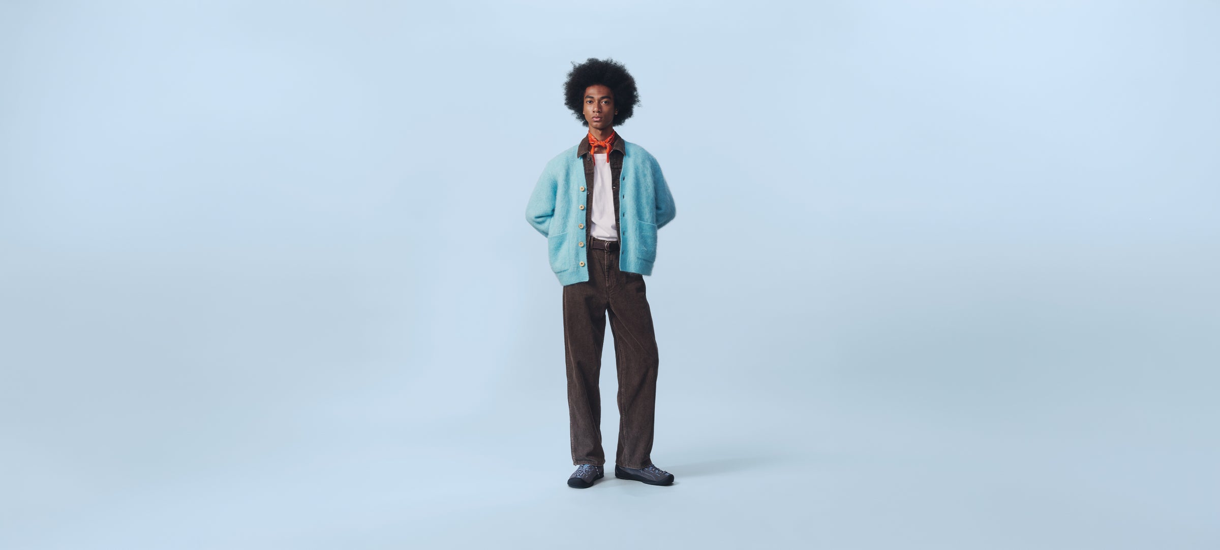 A man with an afro hairstyle stands against a light blue studio background wearing a light blue cardigan, white shirt, orange tie, and dark brown trousers.