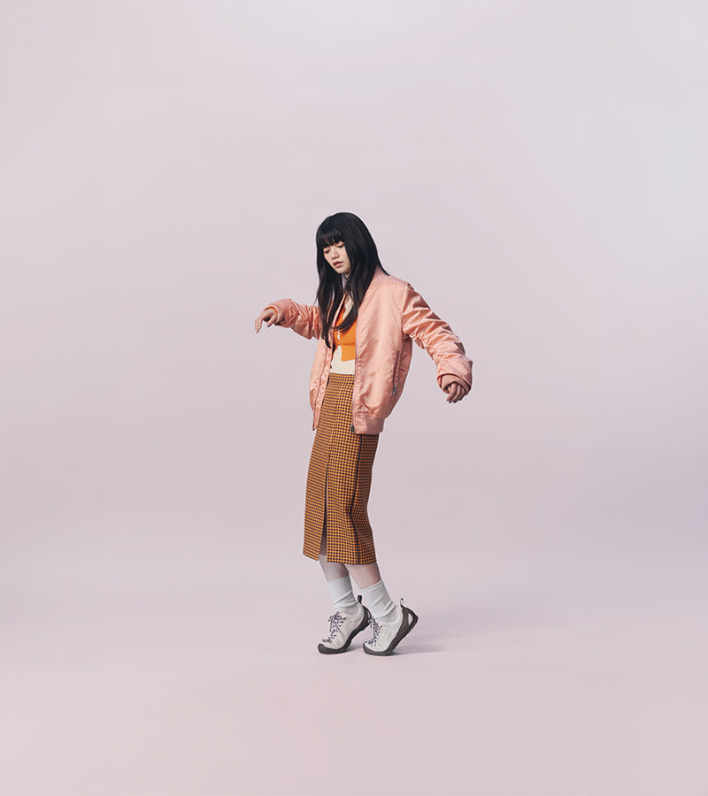 A model with long black hair poses in a studio setting wearing a light pink bomber jacket, a brown and white checkered midi skirt, white socks, and gray sneakers against a solid pale pink background.