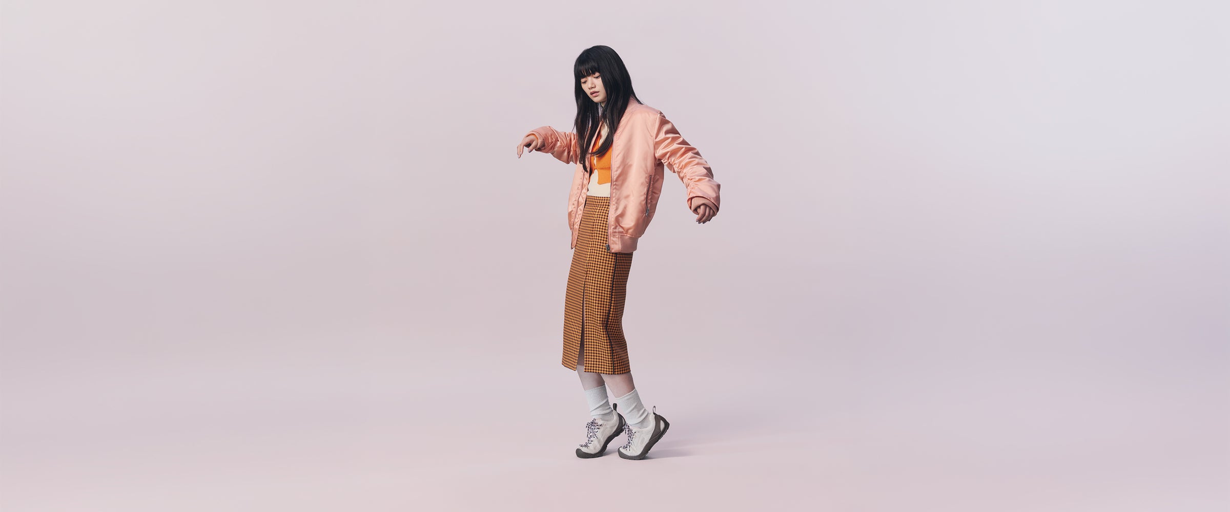 A model with long black hair poses in a studio setting wearing a light pink bomber jacket, a brown midi skirt, a white crop top, white socks, and black and white sneakers against a solid pale pink background.