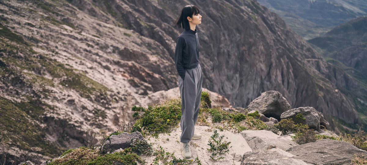 A person standing on a rocky mountain path, wearing a dark long-sleeve top and loose gray pants, with steep rocky mountains in the background.