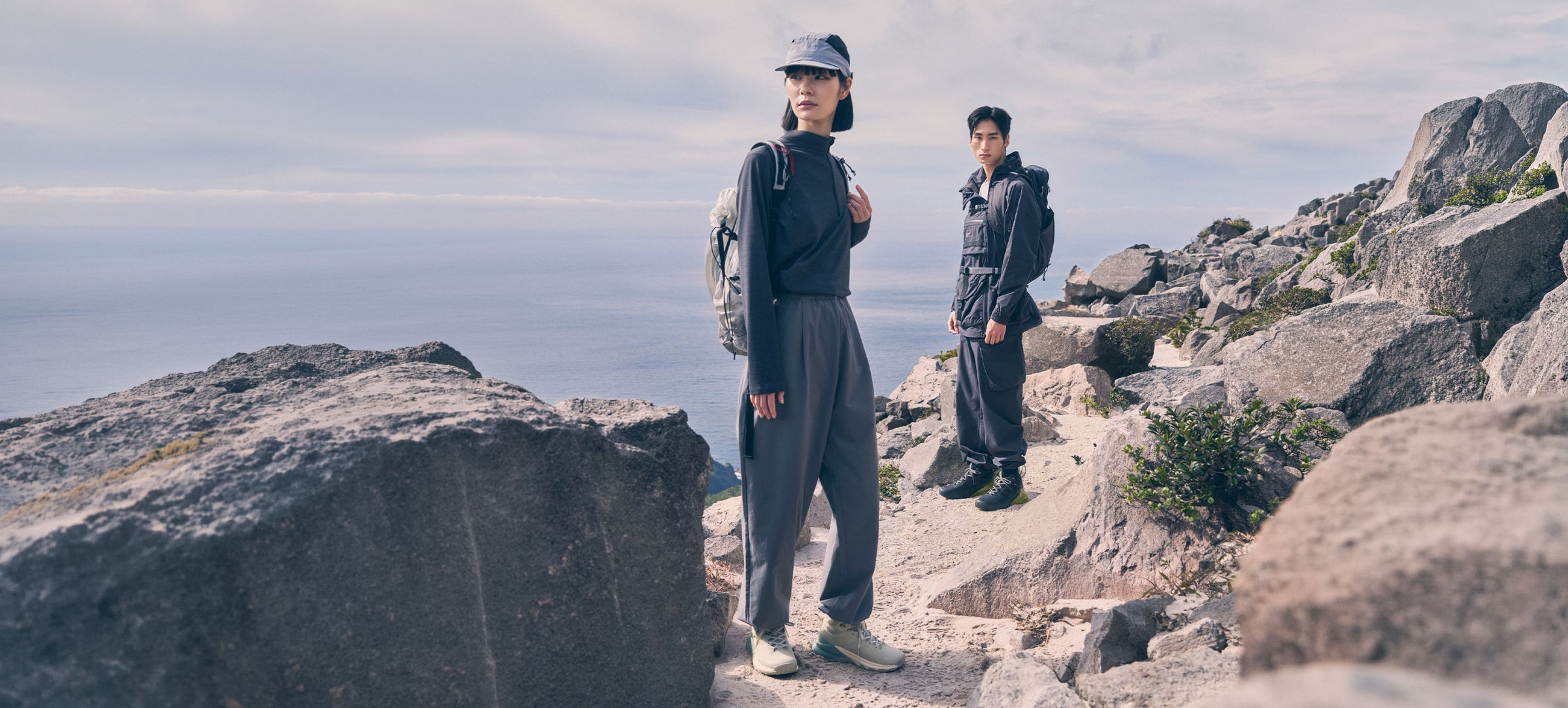 Two people standing on a rocky outdoor trail with a cloudy sky and distant water in the background. The person in front wears a dark cap, dark jacket, loose dark pants, and light-colored shoes, while the second person behind wears a dark jacket and dark pants.
