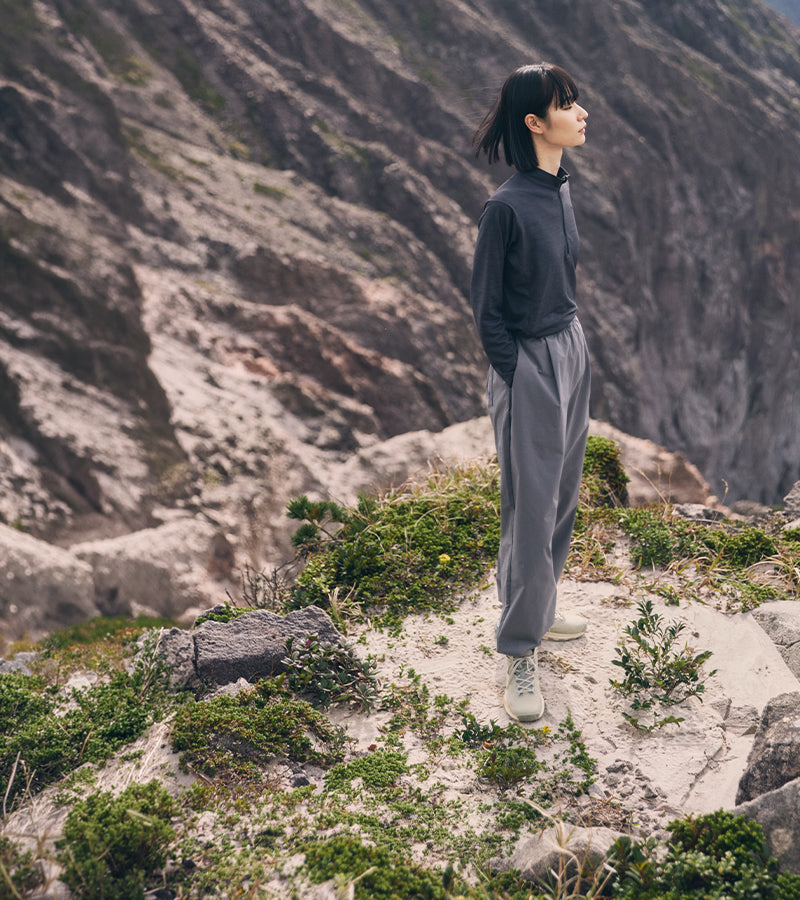 A woman stands on a rocky mountain path wearing a dark gray long-sleeve top and gray sweatpants, with a rugged mountain landscape in the background.