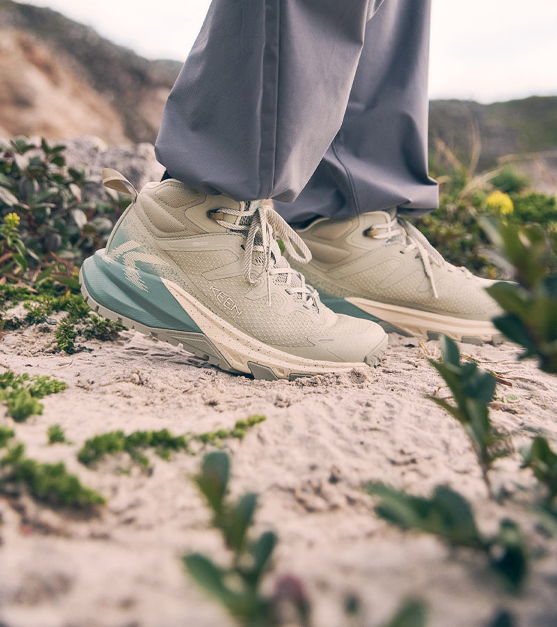 A close-up of a person wearing light beige and mint green KEEN Apex shoes while walking outdoors on sandy ground with small plants and hills in the background.