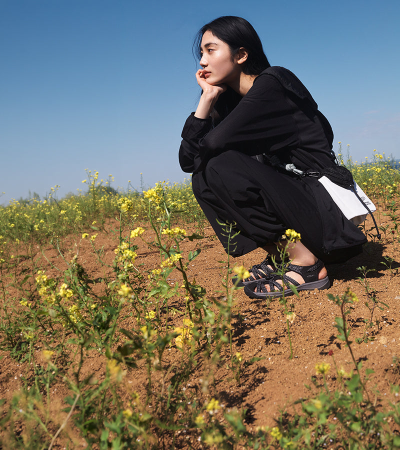 A person in a black outfit crouching outdoors in a field of yellow flowers under a clear blue sky, with one hand near their face and sandals visible on the ground.