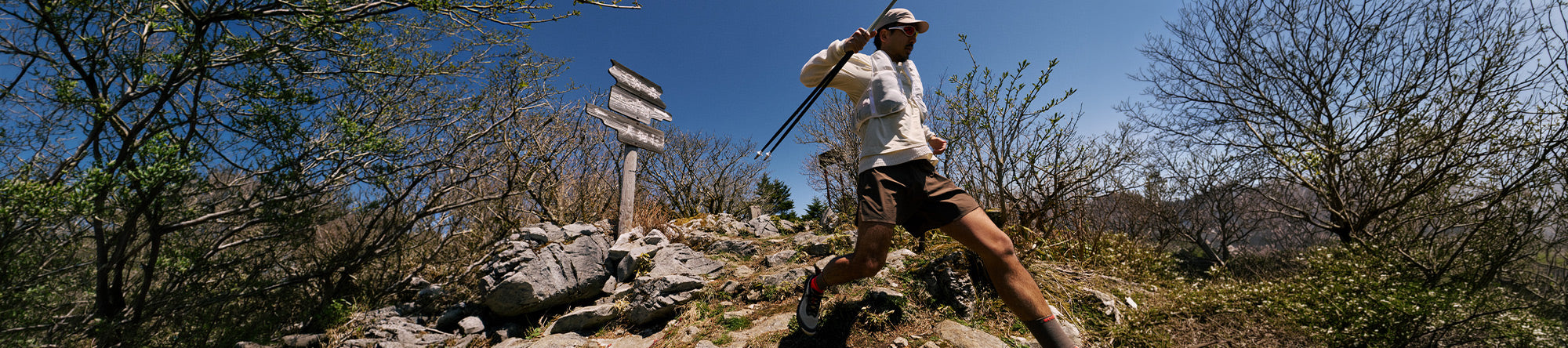 A man hiking outdoors on a rocky trail under a blue sky, wearing a white long-sleeve shirt, brown shorts, and a white cap, while carrying a rifle over his shoulder. Trees and rocks are visible in the background.