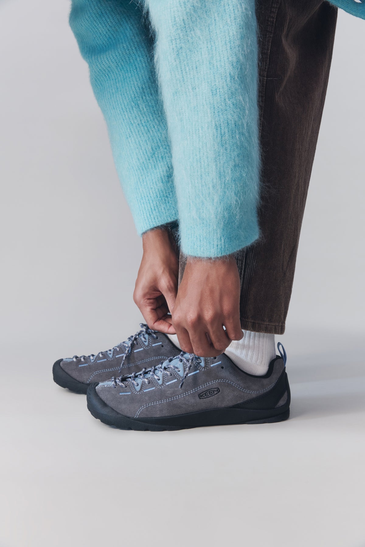 A person wearing gray lace-up shoes with white socks and a turquoise sweater, tying the shoe on the foreground against a light gray studio background.
