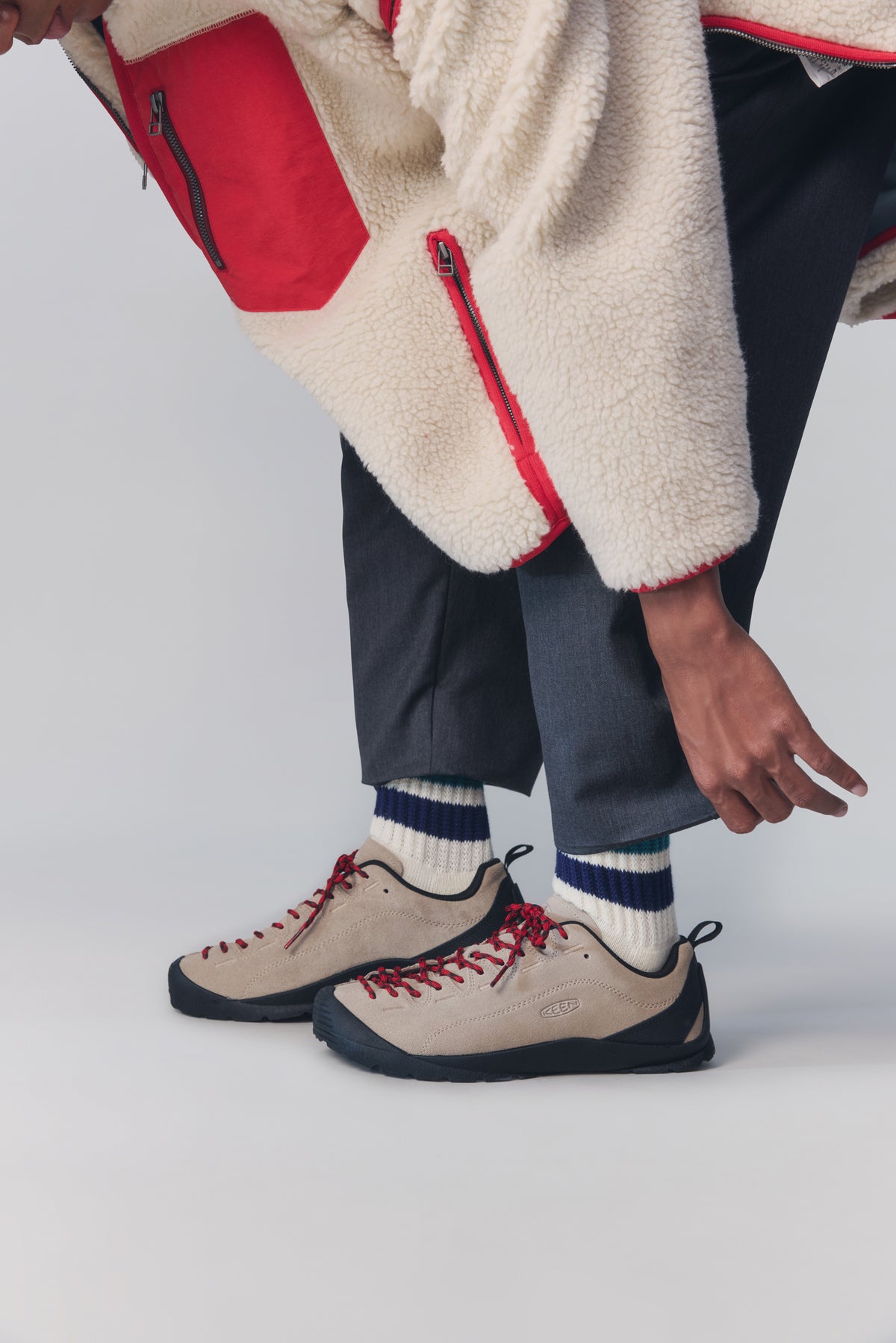 A close-up of a person wearing beige technical climbing shoes with red laces, navy socks, and dark trousers, styled with a cream sherpa fleece jacket with red accents.