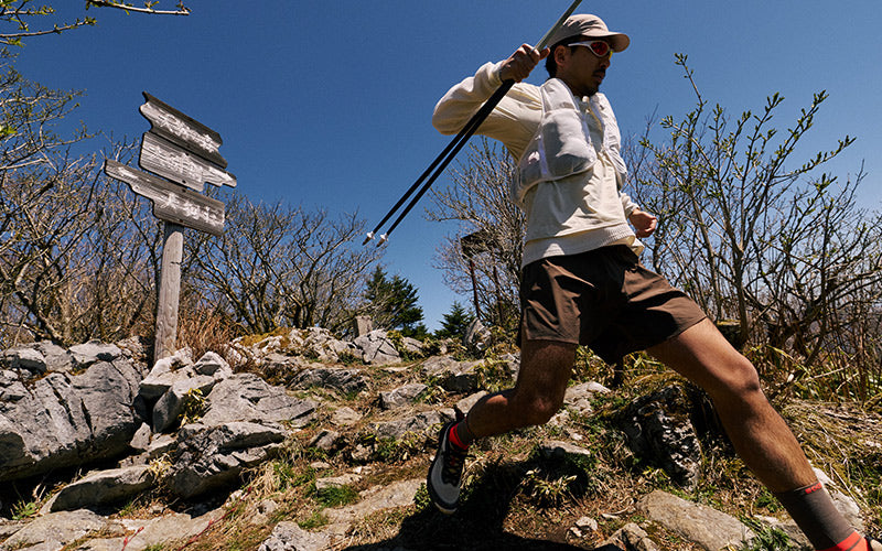 A man trail running uphill on a rocky mountain path under a clear blue sky, carrying trekking poles. He is wearing a light beige long-sleeve performance hoodie, brown shorts, and running shoes.