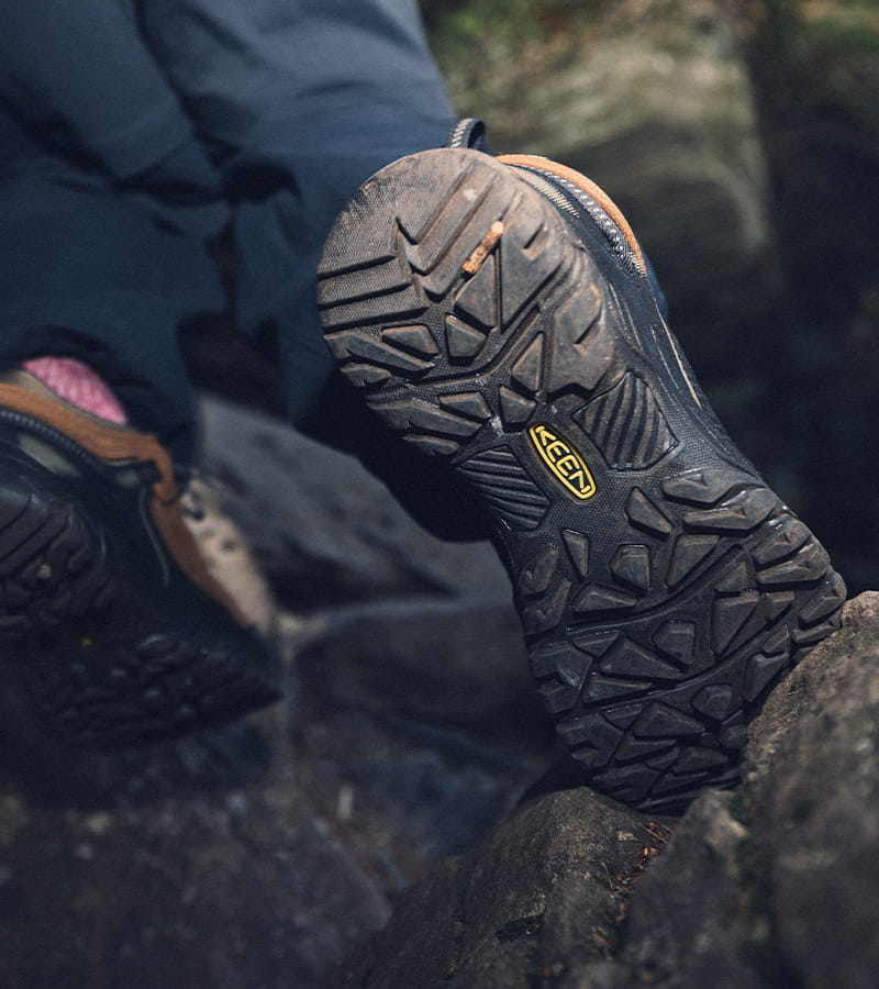 A close-up of a hiking boot's textured black sole with a yellow Keen logo, resting on rocks outdoors with a person's legs visible in the background.