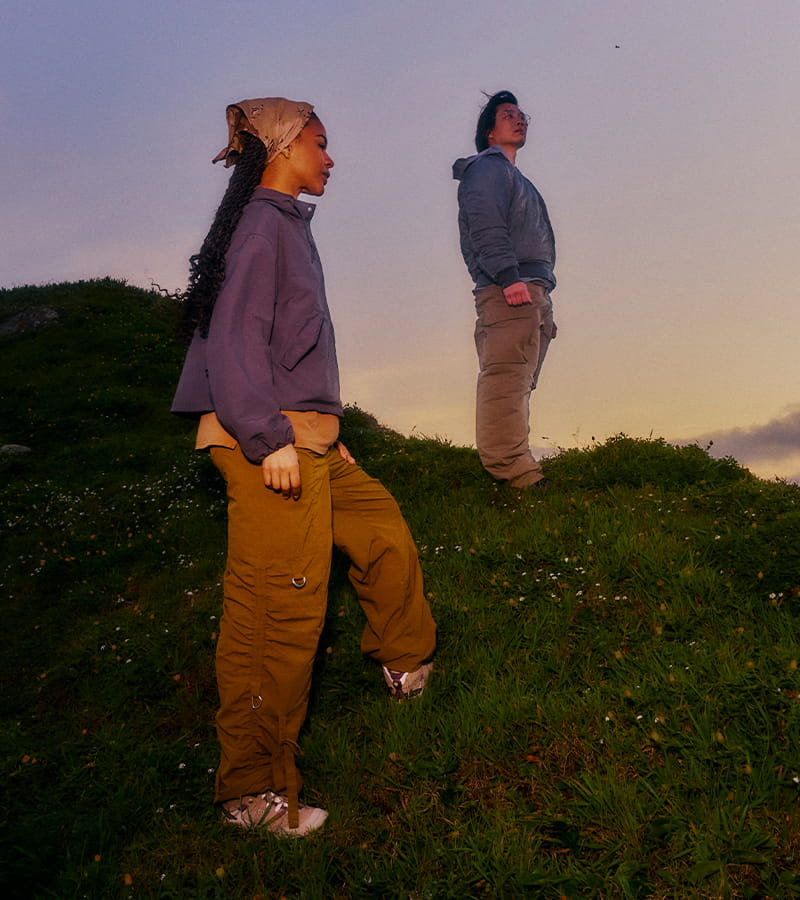 Two people standing on a grassy hill at sunset, wearing oversized streetwear-style outfits. One person in the foreground wears a purple hoodie, tan cargo pants, and a beige headscarf, while the second person behind wears a gray hoodie and beige pants.
