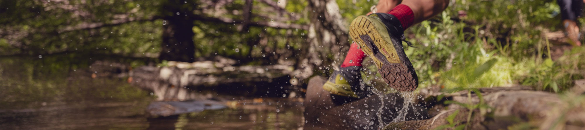 Person wearing rubber boots wading through a stream with trees and greenery in the background
