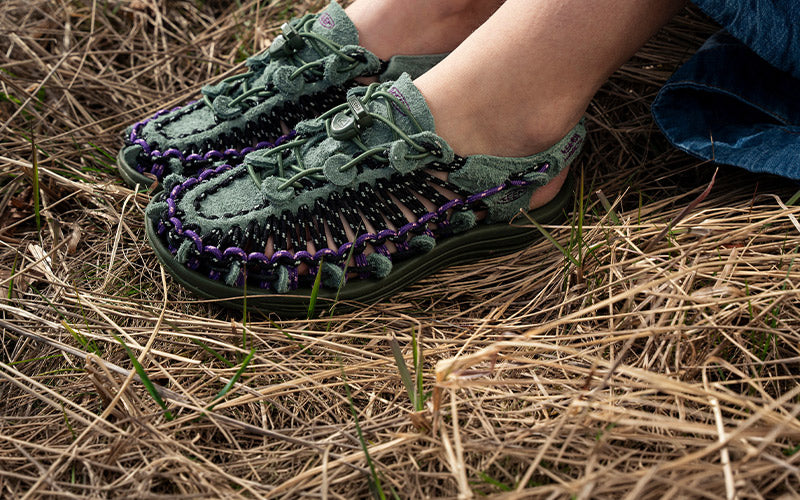 A close-up of a person wearing dark green woven moccasin-style shoes with purple braided trim, standing on dry grass outdoors.