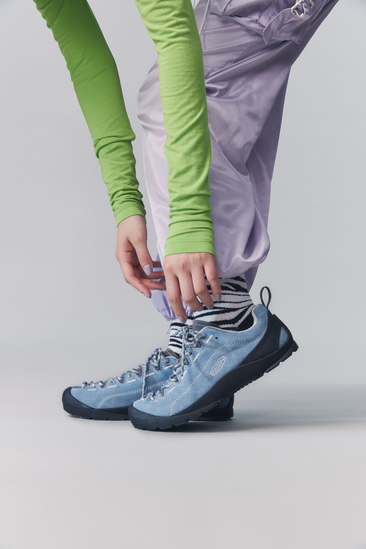 A person wearing light blue shoes with black and white striped socks, shown from the waist down while tying the shoe laces. The background is a clean, neutral gray studio backdrop.